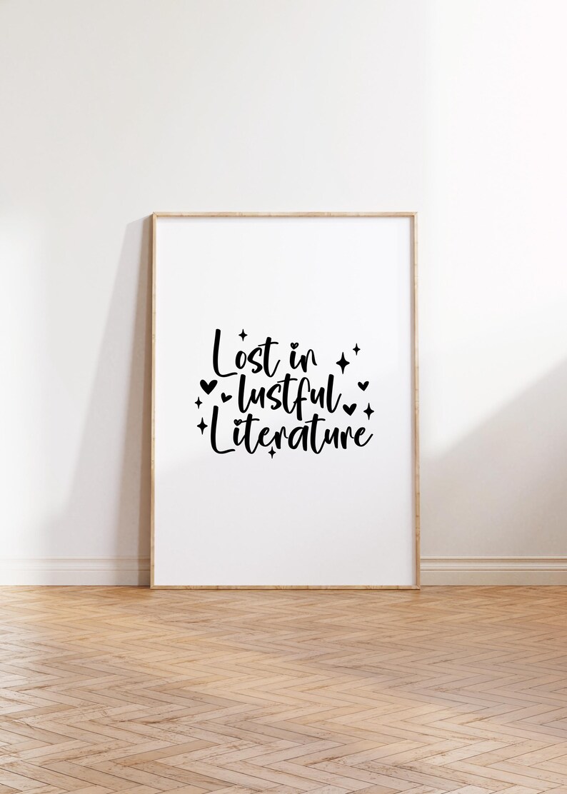Puede incluir: Una impresi&oacute;n enmarcada con el texto "Lost in lustful literature" en escritura negra con peque&ntilde;os corazones y estrellas alrededor de las palabras. La impresi&oacute;n est&aacute; sobre un fondo blanco y est&aacute; enmarcada en un marco de madera claro.