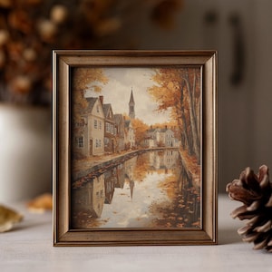 May include: A framed painting depicting a town scene in autumn with houses lining a canal, a church steeple in the distance, and golden trees reflecting in the water. The artwork is enclosed in a simple, metallic-toned rectangular frame.