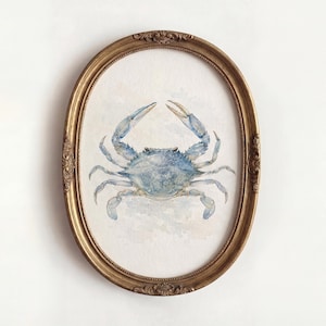 May include: A watercolor illustration of a blue crab centered within an ornate, oval-shaped antique gold frame. The crab is depicted with shades of light and dark blue, featuring detailed claws and legs against a soft, off-white textured background.