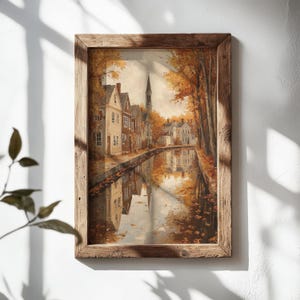 May include: A framed wall art print depicting a canal scene during autumn. The image features houses with light-colored siding, a tall church spire, and trees with orange and yellow foliage. The scene is reflected in the calm water of the canal.