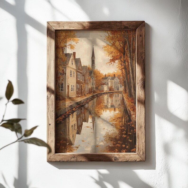 May include: A framed wall art print depicting a canal scene during autumn. The image features houses with light-colored siding, a tall church spire, and trees with orange and yellow foliage. The scene is reflected in the calm water of the canal.