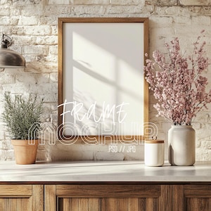 以下が含まれることがあります： 白い背景のウッドフレームのモックアップが、白い表面のカウンタートップの上に置かれています。カウンタートップには、鉢植えの植物とピンクの花の入った花瓶があります。フレームはレンガの壁の背景にあります。