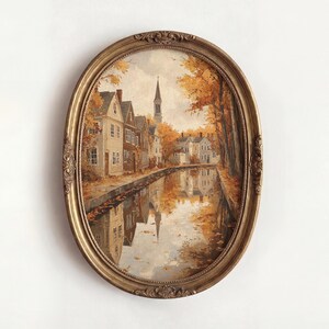 May include: An oval-shaped painting in an ornate gold frame depicting a canal scene during autumn. The artwork features houses along the water, golden-hued trees, and a church steeple in the distance, with reflections visible on the water surface.