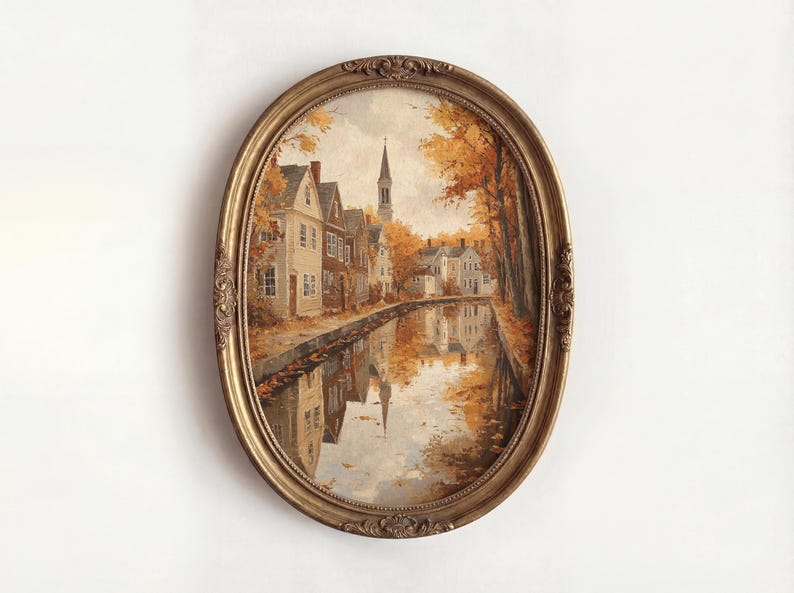 May include: An oval-shaped painting in an ornate gold frame depicting a canal scene during autumn. The artwork features houses along the water, golden-hued trees, and a church steeple in the distance, with reflections visible on the water surface.