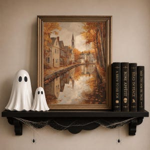 May include: A framed landscape painting of a town along a canal during autumn, displayed on a black shelf with two white ceramic ghost figurines and four books titled A Brew to Die For, Bone Appetit, Read in Peace, and May Thy Aim Be True.