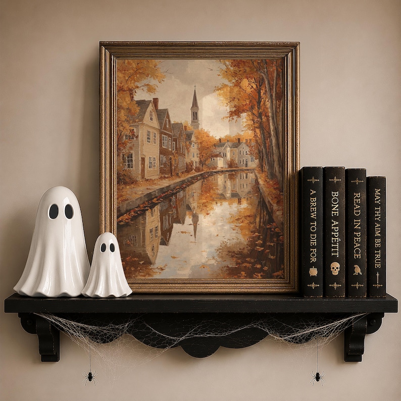 May include: A framed landscape painting of a town along a canal during autumn, displayed on a black shelf with two white ceramic ghost figurines and four books titled A Brew to Die For, Bone Appetit, Read in Peace, and May Thy Aim Be True.