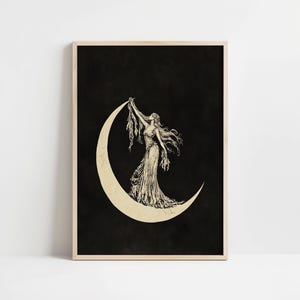 May include: A framed black and white print featuring a woman standing on a crescent moon. The woman is wearing a long dress and holding a flowing fabric. The background is black, and the moon is a light beige color.