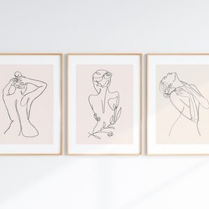 May include: Three framed art prints featuring minimalist line drawings of the female form. Each print has a beige background and a thin, black line drawing. The frames are a light wood color, and the art is displayed on a white wall.