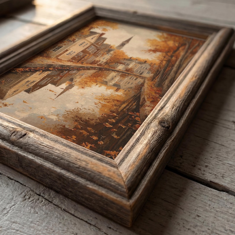 May include: A rectangular painting in a weathered, dark brown wooden frame. The artwork depicts a landscape scene with buildings, a bridge, and trees in shades of orange, brown, and cream, reflecting in a body of water during the autumn season.