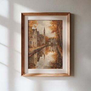 May include: A framed wall art print depicting a canal scene in autumn with golden trees, historic houses, and a church steeple. The water reflects the buildings and foliage, creating a symmetrical composition within a light wood frame and mat.