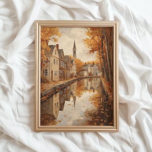 May include: A framed wall art print depicting a canal scene in autumn with golden trees, historic houses, and a church steeple. The water reflects the buildings and foliage in warm orange, brown, and yellow tones, all encased in a light gold frame.