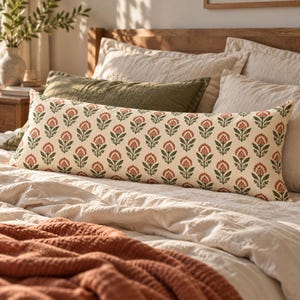 May include: A long rectangular lumbar pillow featuring an off-white base color with a repeating pattern of stylized orange flowers and green leaves. The pillow is positioned horizontally across a bed with other pillows and a textured blanket.