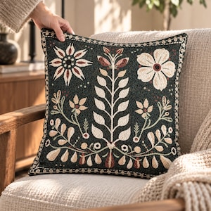 May include: A square throw pillow featuring a dark green fabric base with a woven cream and reddish-brown floral and botanical pattern. The design includes a central vertical stem with leaves, surrounded by various flowers and foliage.