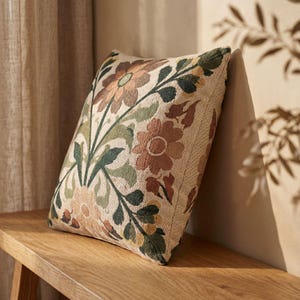 May include: A square throw pillow featuring a woven floral pattern in shades of terracotta, sage green, and cream. The design displays large stylized flowers and leafy stems across the front surface, creating a textured and detailed botanical appearance.