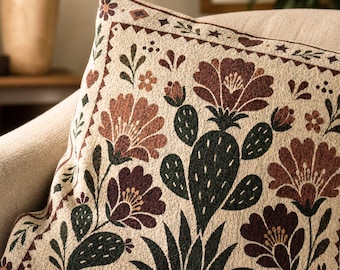 May include: A square throw pillow featuring a woven cream-colored fabric with a repeating pattern of dark green cacti and deep red flowers. The design includes a decorative border with small geometric shapes, leaves, and stars framing the central floral motif.