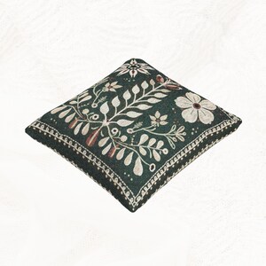 May include: A square throw pillow featuring a dark green background with a cream-colored botanical pattern of leaves and flowers. The design includes a decorative border with a repeating stitch-like pattern along the edges of the pillow.