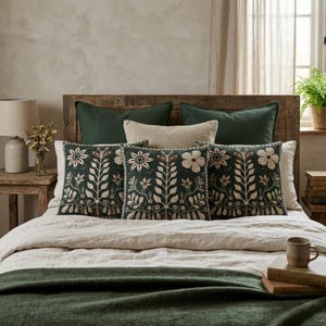 May include: Three square throw pillows featuring a dark green background with a symmetrical cream and brown floral and botanical embroidery pattern. The pillows are arranged on a bed with a light beige duvet and a dark green throw blanket.