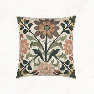 May include: A square throw pillow featuring a woven tapestry design with a central floral motif. The pattern displays large brown, peach, and light green flowers surrounded by dark green leaves and stems against a light beige textured background.