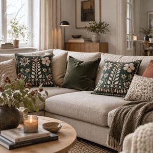 May include: A beige sectional sofa featuring two square throw pillows with dark green fabric and cream-colored floral embroidery, a solid olive green velvet pillow, a striped pillow, and a small floral print pillow with a textured throw blanket.
