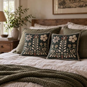 May include: Two square throw pillows featuring dark green fabric with cream and rust-colored floral and botanical embroidery. The pillows have a decorative stitched border and are placed on a bed with a light-colored duvet and green blanket.
