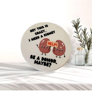 May include: White round sticker with a cartoon illustration of two red kidneys holding a sign that says "HELP!" The text on the sticker reads "Hey this is crazy... I need a kidney. Be a donor, maybe?"