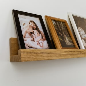 Può includere: Una mensola galleggiante in legno che espone tre fotografie incorniciate. Le cornici sono nere, marroni e bianche, contenenti ritratti di famiglia e una foto di matrimonio. La mensola è in legno chiaro ed è montata su una parete bianca.