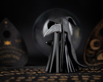 May include: A black Grim Reaper figurine with a silver scythe stands before a blurred glass orb. Two Ouija boards flank the figure, set against a dark background. The scene evokes a sense of mystery and the supernatural.