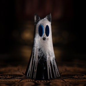 May include: A white ghost figurine with blue eyes and a black base. The ghost is standing on a patterned surface.