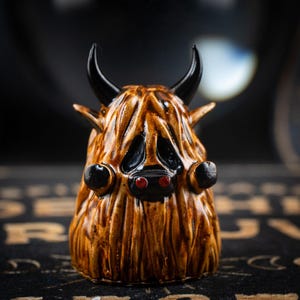 May include: A brown ceramic figurine of a hairy creature with black horns and red eyes. The creature has a black nose and a furry face.