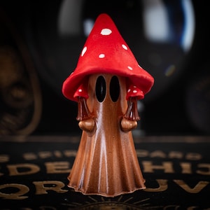 May include: A whimsical, brown mushroom-shaped figurine with a red cap dotted with white spots. The figurine has two small mushroom accents and two large, oval-shaped eye holes. The background is dark and blurred.