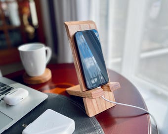May include: A wooden phone stand with a black smartphone resting on it. The stand is on a wooden table with a white laptop and a white charging block.