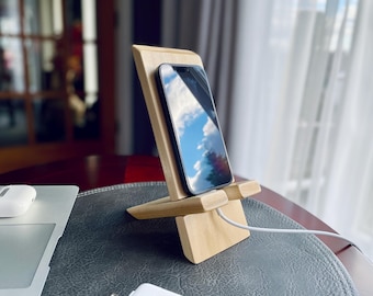 May include: A wooden phone stand with a smartphone resting on it. The stand is light brown and has a simple design. The phone is black and has a blue sky reflected on the screen.