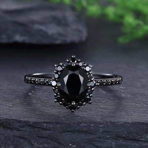 Op de afbeelding: Een zwarte edelsteenring met een ovaalvormige centrale steen en een halo van kleinere stenen. De band is ook versierd met kleine zwarte stenen. De ring is geplaatst tegen een donkere achtergrond, wat het elegante ontwerp benadrukt.