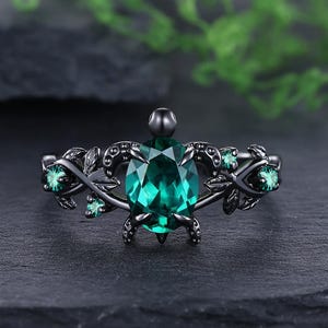 May include: Black metal ring featuring a large oval-cut emerald green gemstone as the centerpiece. The ring is adorned with smaller green gemstones and intricate leaf-like designs on the band, creating an elegant and nature-inspired aesthetic.