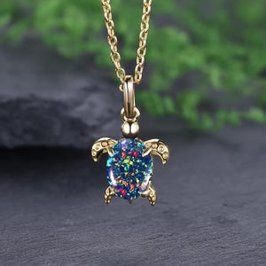 Op de afbeelding: Een goudkleurige ketting met een schildpadhanger. De hanger heeft een donkerblauwe, ovaalvormige kern met kleurrijke vlekken en is omlijst door een goudkleurig schildpadontwerp. De ketting is ook goudkleurig.
