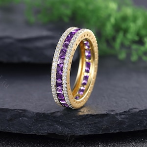 May include: A gold ring featuring two rows of square-cut purple gemstones and a single row of small white gemstones. The ring is set against a dark background with green foliage.
