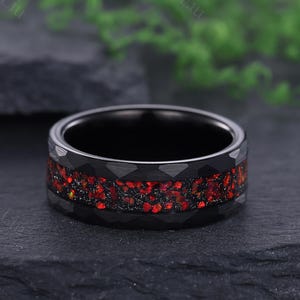 Puede incluir: Un anillo negro con una textura martillada y una banda de piedras preciosas rojas. El anillo se encuentra sobre un fondo oscuro, mostrando el contraste entre el metal negro y las vibrantes piedras rojas. Una pieza de joyería moderna.