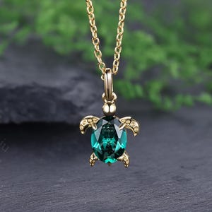 May include: A gold-colored necklace with a pendant shaped like a turtle. The turtle pendant features a large, oval-shaped, emerald-green gemstone. The turtle's flippers and head are detailed with small, gold-colored accents. The necklace chain is also gold-colored.