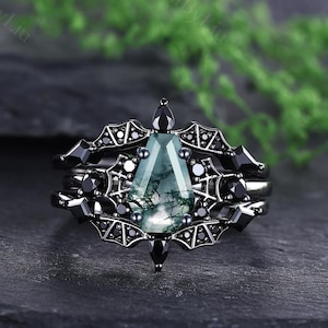 May include: A black metal ring featuring a coffin-shaped moss agate centre stone with green and black inclusions. The band is designed with intricate bat wing motifs and small black accent stones surrounding the central gemstone setting.