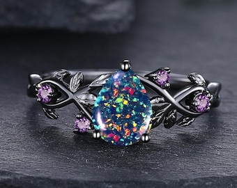 May include: Black ring featuring a pear-shaped, iridescent opal gemstone. The ring has a floral design with leaves and small purple gemstones. The ring is set on a dark surface.