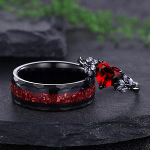 May include: Two rings. One is black with a red inlay and a hammered texture. The other is black with a heart-shaped red stone and white accents. The rings are displayed on a dark gray surface.