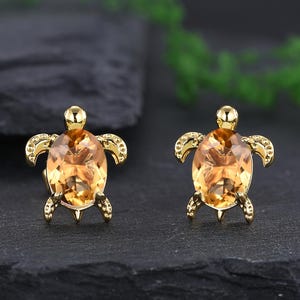 Può includere: Un paio di orecchini a lobo a forma di tartaruga dorata. Ogni orecchino presenta una grande pietra preziosa ovale color ambra incastonata in una montatura in metallo dorato. Il design della tartaruga include gambe, pinne e testa dettagliate.