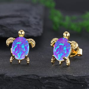 May include: A pair of gold turtle earrings with iridescent purple opal stones. The earrings are small and delicate, perfect for everyday wear.