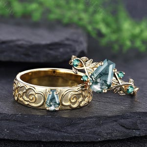 Pode incluir: Um conjunto de anéis de ouro com uma banda de motivos de nuvens em relevo e uma pedra de ágata musgo em forma de caixão, combinada com um anel com uma ágata musgo central, pequenas pedras verdes e detalhes metálicos em forma de folha.