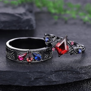 May include: Two black rings. One ring features a band with a leaf pattern and three colored gemstones: pink, red, and blue. The other ring has a large red gemstone and smaller pink and blue gemstones, with a floral design.