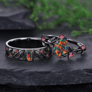 May include: Two black opal rings. One ring features an oval-shaped opal with a floral design and red accents. The other ring has a rectangular opal and a floral design with red accents. The rings are set against a dark background.