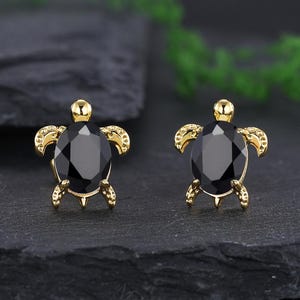 Può includere: Un paio di orecchini a lobo a forma di tartaruga color oro, ciascuno con una grande gemma nera a taglio ovale. Gli orecchini presentano dettagliati disegni di tartarughe con piccoli piedi e un guscio strutturato. Gli orecchini sono esposti su una superficie scura e strutturata.