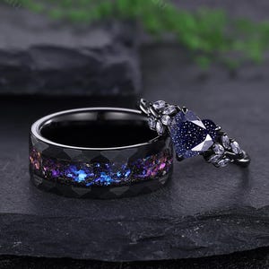 May include: Two black rings. One ring has a galaxy-like inlay with blue, purple, and pink hues. The other ring features a dark blue, heart-shaped stone and small white stones. The rings are set against a dark background.
