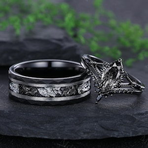 May include: A black wedding ring set. One ring has a hammered texture with a band of silver-colored stone. The other ring has a black gemstone with black accents. The rings are displayed on a dark surface.