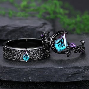 May include: Two black rings with intricate designs. One ring features a central teal-colored gemstone and a floral design. The other ring has a teal gemstone and a crescent moon shape with purple accents. The rings are set against a dark background.